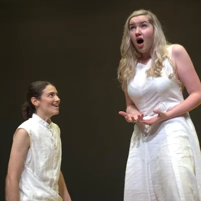 Two young women dressed in white, one has her mouth open as if to sing, with effort. 
