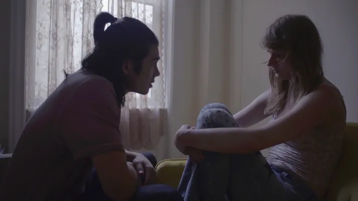 A woman sits on a couch, clutching her knees to her chest, while a man sits in front of her, leaning in to speak