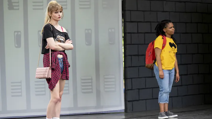 Two young women dressed like teens stand on a stage made to look like a school hallway