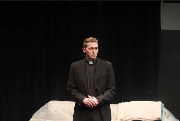 A young man dressed as a priest stands on a sparsely decorated stage