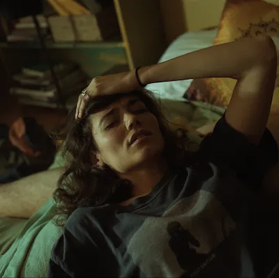 A young woman lies across a bed, her head on the back of a naked man. She clutches her head in pain. 