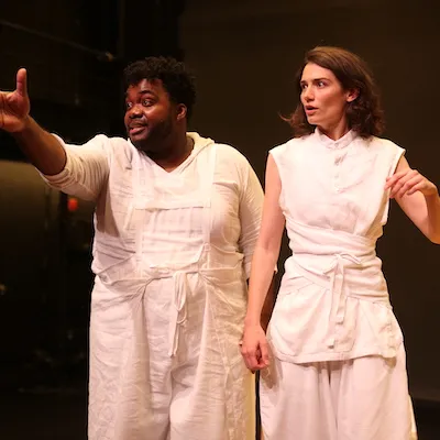 A young man and woman wearing white stand on a stage and signal off toward the left