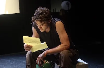 A young man sits on a box on a stage, reading from a yellow piece of paper.