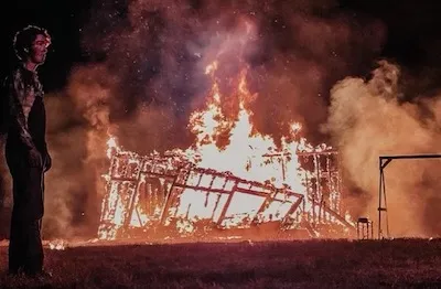 A young man stands in front of a burning building