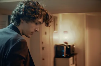A shot of a young man from the side, looking down. He stands in an apartment lit by yellow light. 