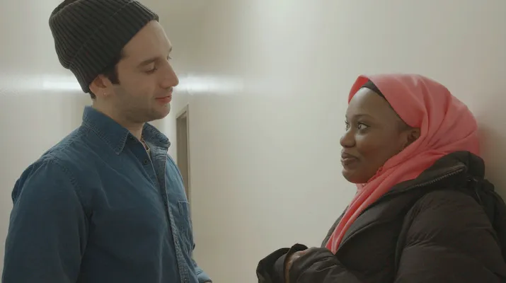 A young woman in a hijab leans against a wall, smiling at a man standing across from her