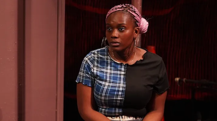 A young woman sits on a box on a stage, expression serious.